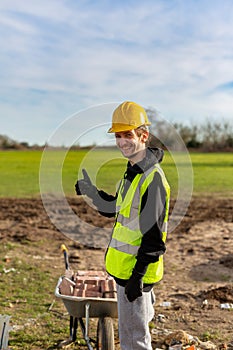 A young adult male builder wearing a high visibility vest and hard hat while giving a thumbs up
