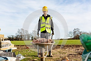 A young adult male builder wearing a high visibility vest and hard hat pushing a wheelbarrow full of bricks while on a building