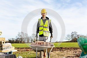 A young adult male builder wearing a high visibility vest and hard hat pushing a wheelbarrow full of bricks while on a building