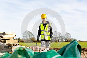 A young adult male builder wearing a high visibility vest and hard hat pushing a wheelbarrow full of bricks while on a building