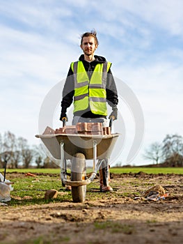 A young adult male builder wearing a high visibility vest and hard hat pushing a wheelbarrow full of bricks while on a building