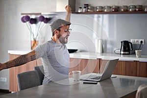 Young adult man with beard and tattoo yawns, stretching in front of laptop on table with cup of coffee  and cell phone. lifestyle