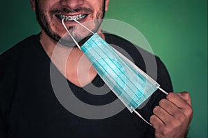 A young adult man with a black T-shirt Biting a light blue disposable mask with light blue folds with white strips on a sea green