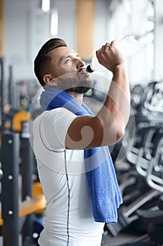 Young adult man drinking bottle of water on trreadmill in gym.