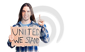 Young adult man with long hair holding united we stand banner surprised with an idea or question pointing finger with happy face,
