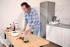 Young adult man preparing salad, chopping vegetables on a cutting board in white interior of modern minimalist kitchen