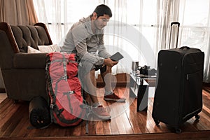 Young adult man sitting in the armchair at home checking his passport near his backpack and travel suitcase in the living room of