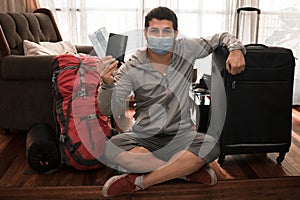 Young adult man sitting on the wooden floor with his backpack and holding his travel suitcase and his passport with disposable mas