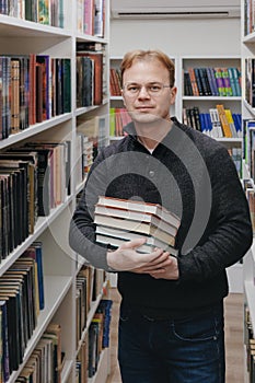 young adult man stands in library in middle of the shelves and holds stack of books in hands.