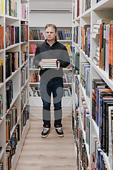 young adult man stands in library in middle of the shelves and holds stack of books in hands.