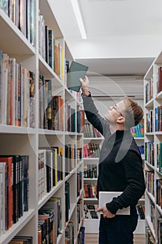 young adult man stands in library in middle of shelves and takes book. reading and recreation