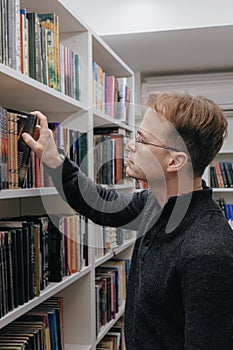 young adult man stands in library in middle of shelves and takes book. reading and recreation