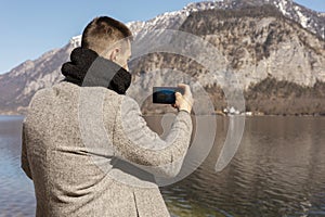 Young adult man taking picture with his smartphone, enjoying mountains, lake, good weather, blue sky, sun. Beautiful
