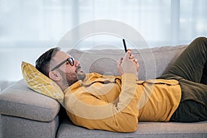 Young adult mature man using smart phone resting lying down on sofa couch at home messaging on mobile phone Guy with eyeglasses