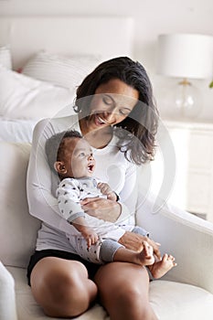 Young adult mother sitting in an armchair in her bedroom, holding her three month old baby son in her arms and looking down at him