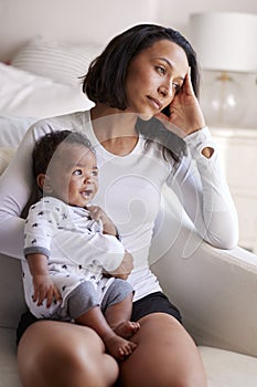 Young adult mother sitting in an armchair in her bedroom, holding her three month old baby son, looking away in contemplation, ver