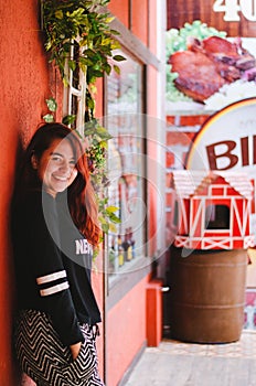 A young adult redhead brazilian smiling to the camera while posing next to a shop display with red walls and a trash bin shaped as