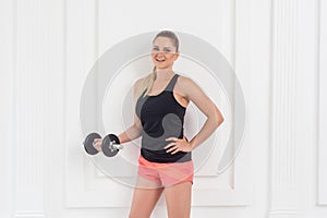 Young adult satisfied athletic beautiful bodybuilder woman in pink shorts and black top standing, holding dumbells doing exercise