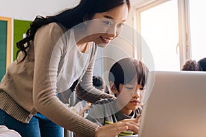 Young adult smiling beautiful Asian teacher helping elementary student boy with laptop in computer classroom