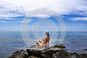 Young adult woman in bikini sits on a granite stone and looks at the sea - view from the back. European female tourist sits on a