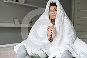 Young adult woman forty years plus size body positive in white blanket with festive cupcake with candle in hands on bed at home