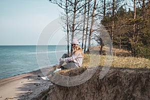 Young adult woman in pink hat sitting alone on the bluffs, looking of sea, freedom concept, peaceful atmosphere