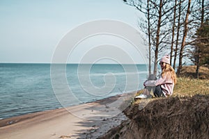 Young adult woman in pink hat sitting alone on the bluffs, looking of sea, freedom concept, peaceful atmosphere