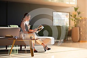 Young adult woman reading magazine on sofa in light modern interior of hotel reception, office building hall, waiting area