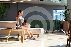 Young adult woman sitting on white sofa in light modern interior of hotel reception, office building hall, waiting area background