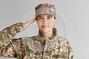 Young adult woman soldier saluting with hand near head, looking at camera with pleasant facial experession, wearing military