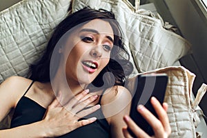 young adult woman surprised using mobile phone while lying in bed