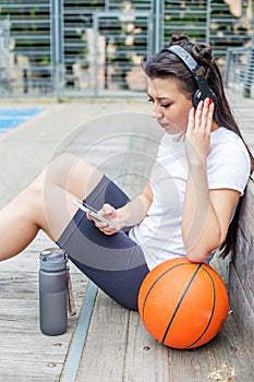 Young adult woman uses wireless headphones and smartphone while sitting on basketball court