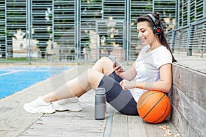 Young adult woman uses wireless headphones and smartphone while sitting on basketball court