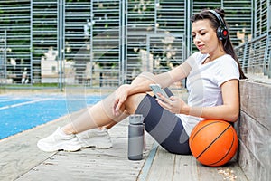 Young adult woman uses wireless headphones and smartphone while sitting on basketball court