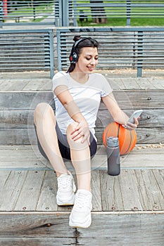 Young adult woman uses wireless headphones and smartphone while sitting on basketball court