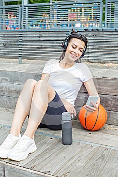 Young adult woman uses wireless headphones and smartphone while sitting on basketball court