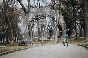 A young adult woman in winter clothing rides her bike along a tranquil park pathway, surrounded by bare trees and nature