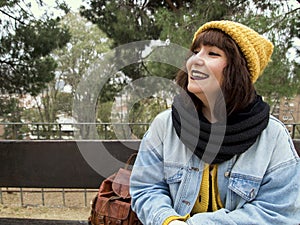 Young adult woman with yellow woolen cap sitting on a park bench and smiling to someone