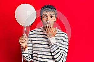 Young african amercian man holding balloon covering mouth with hand, shocked and afraid for mistake