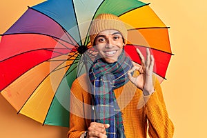 Young african amercian man holding colorful umbrella doing ok sign with fingers, smiling friendly gesturing excellent symbol