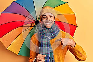 Young african amercian man holding colorful umbrella pointing finger to one self smiling happy and proud