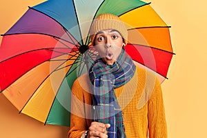 Young african amercian man holding colorful umbrella scared and amazed with open mouth for surprise, disbelief face