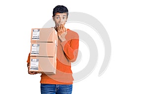 Young african amercian man holding delivery package covering mouth with hand, shocked and afraid for mistake