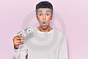 Young african amercian man holding dollars scared and amazed with open mouth for surprise, disbelief face