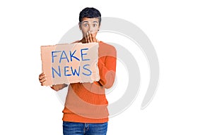 Young african amercian man holding fake news banner covering mouth with hand, shocked and afraid for mistake