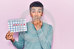 Young african amercian man holding heart calendar covering mouth with hand, shocked and afraid for mistake
