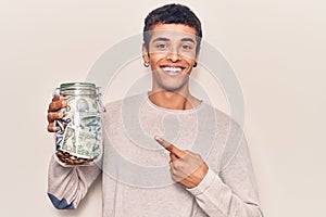 Young african amercian man holding jar with savings smiling happy pointing with hand and finger
