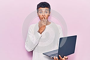 Young african amercian man holding laptop covering mouth with hand, shocked and afraid for mistake