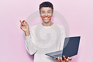 Young african amercian man holding laptop smiling happy pointing with hand and finger to the side