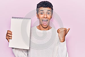 Young african amercian man holding notebook pointing thumb up to the side smiling happy with open mouth